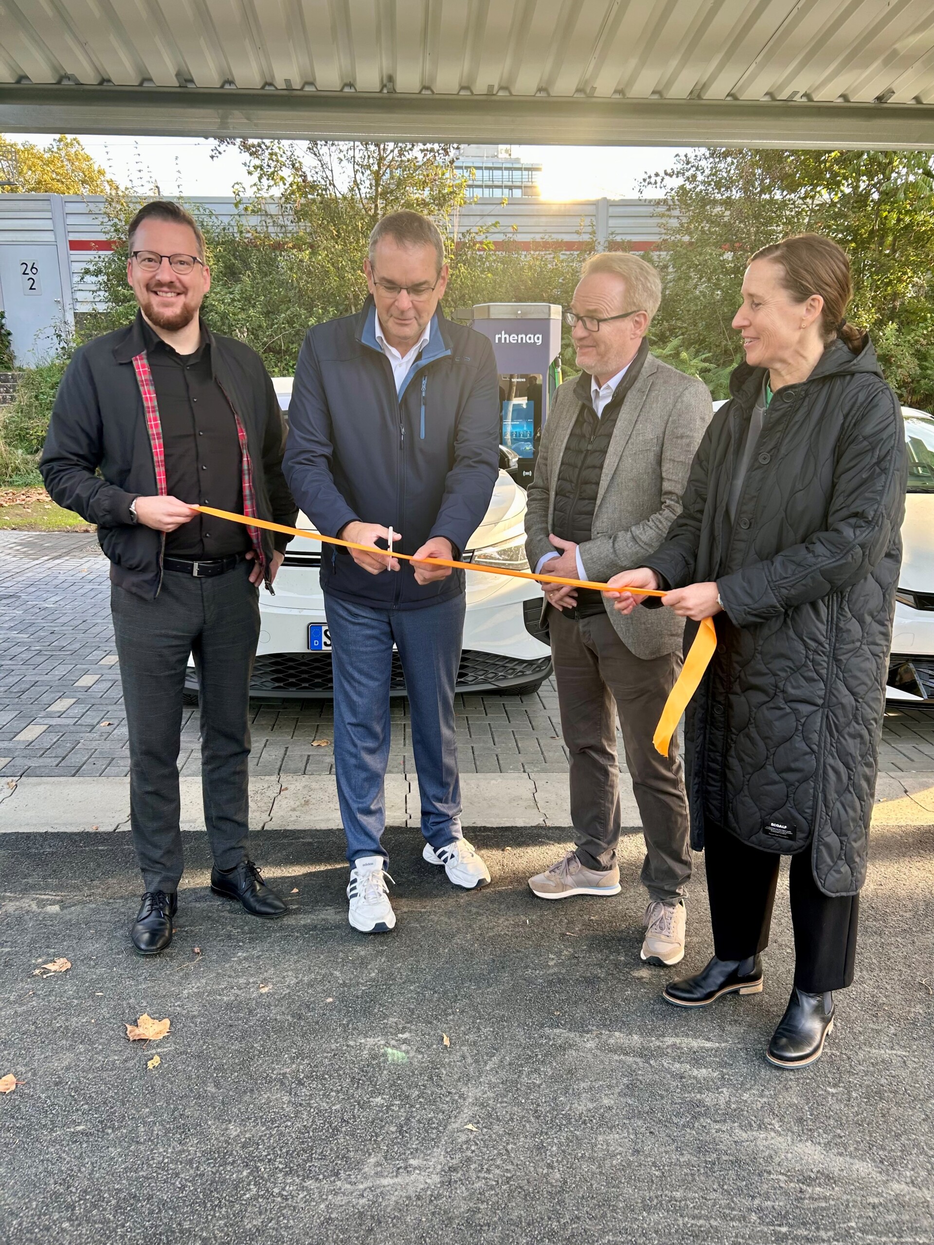 Eröffnung eines Ladeparks: Vier Personen durchtrennen ein orangefarbenes Band vor Elektroautos und einer rhenag-Ladesäule.