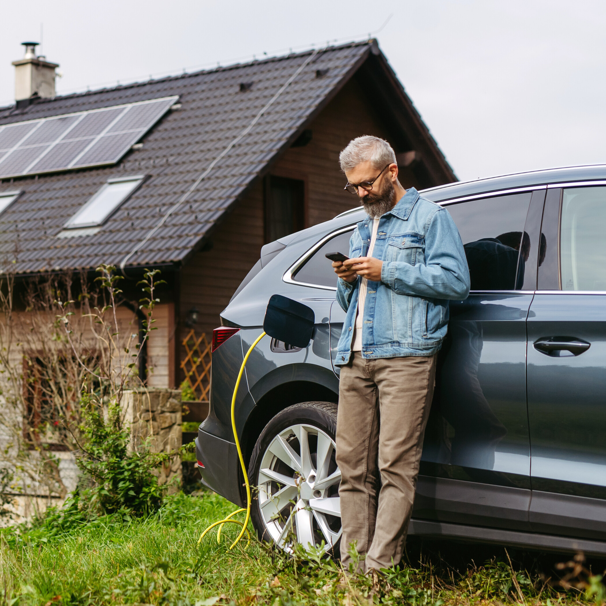 Mann mit einem Handy lehnt an seinem Elektroauto, das gerade vor seinem Haus steht und aufgeladen wird.