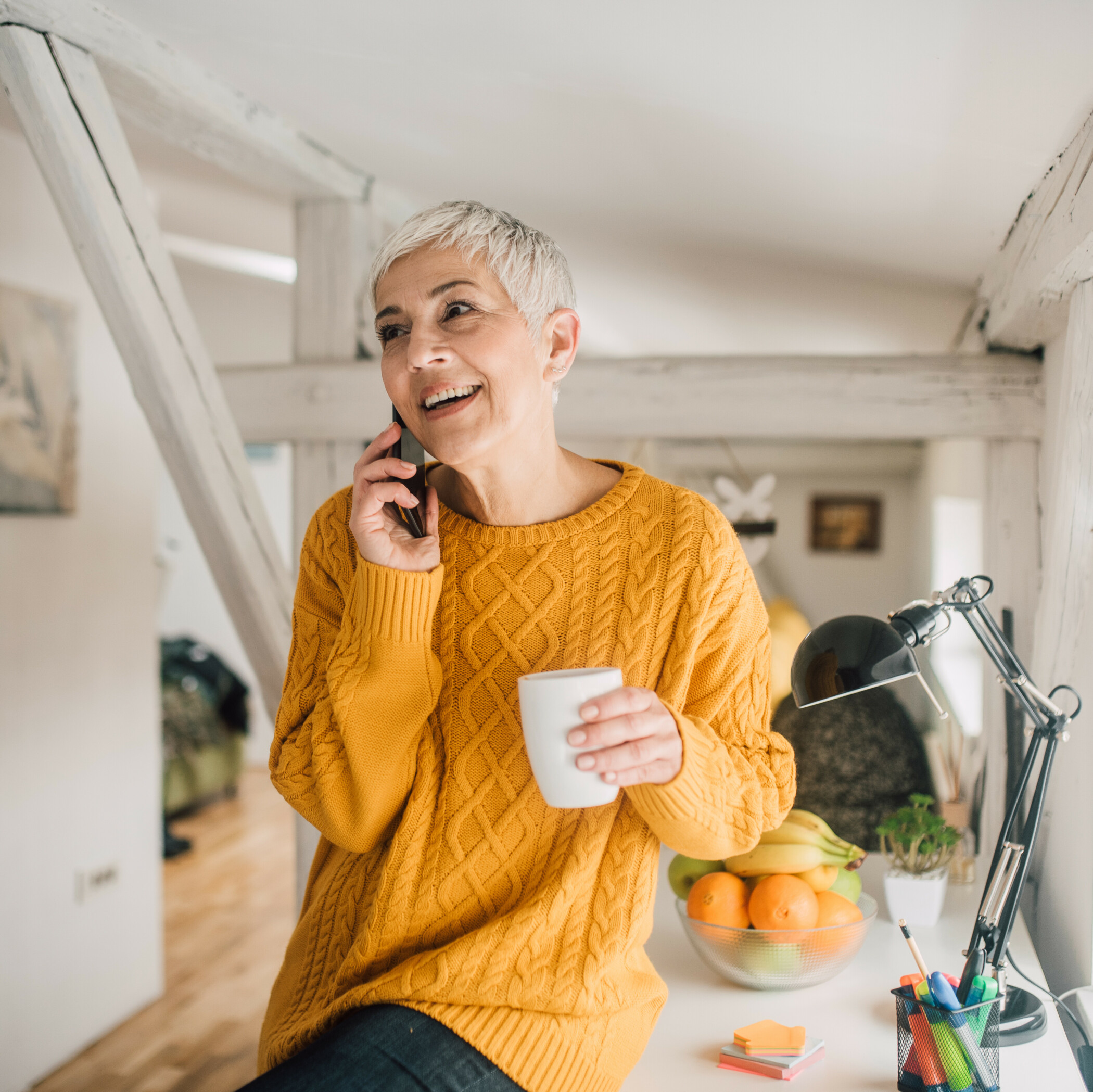 Ältere Frau im orangenen Pulli mit Tasse in der Hand telefoniert lächelnd.