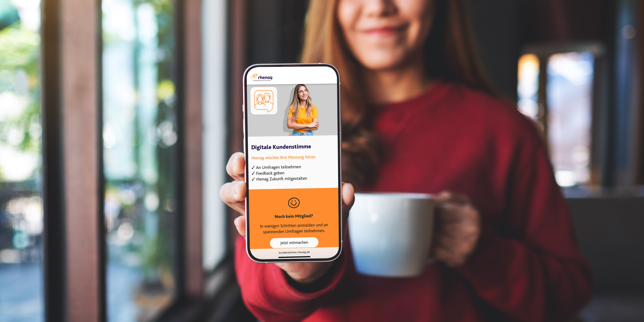 Lächelnde Frau mit rotem Pullover und einer Tasse in der Hand zeigt ihr Handy mit der Website der digitalen Kundenstimme.