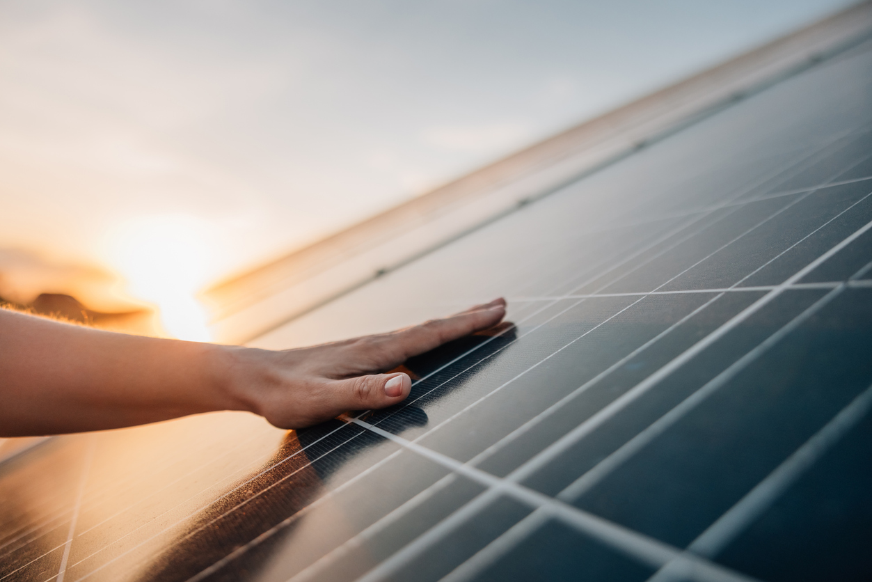 Eine Hand liegt auf einem Solarmodul. Im Hintergrund steht die Sonne tief am Himmel.