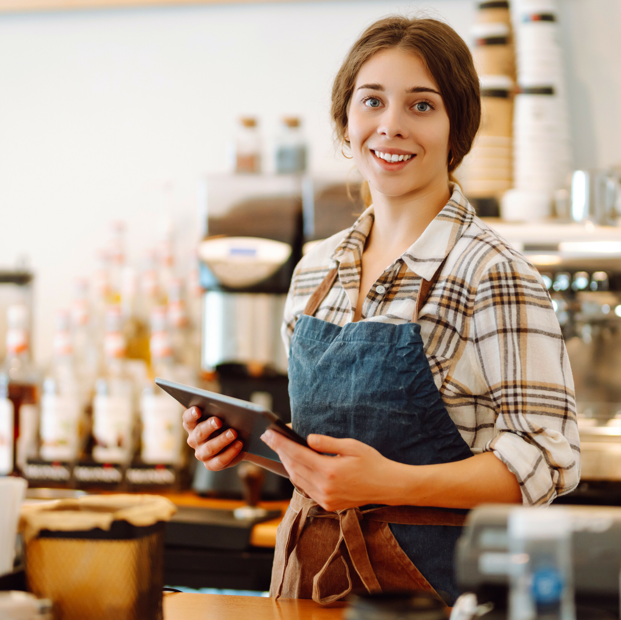Junge Frau mit Schürze und Tablet in der Hand steht in ihrem Café und lächelt in die Kamera.
