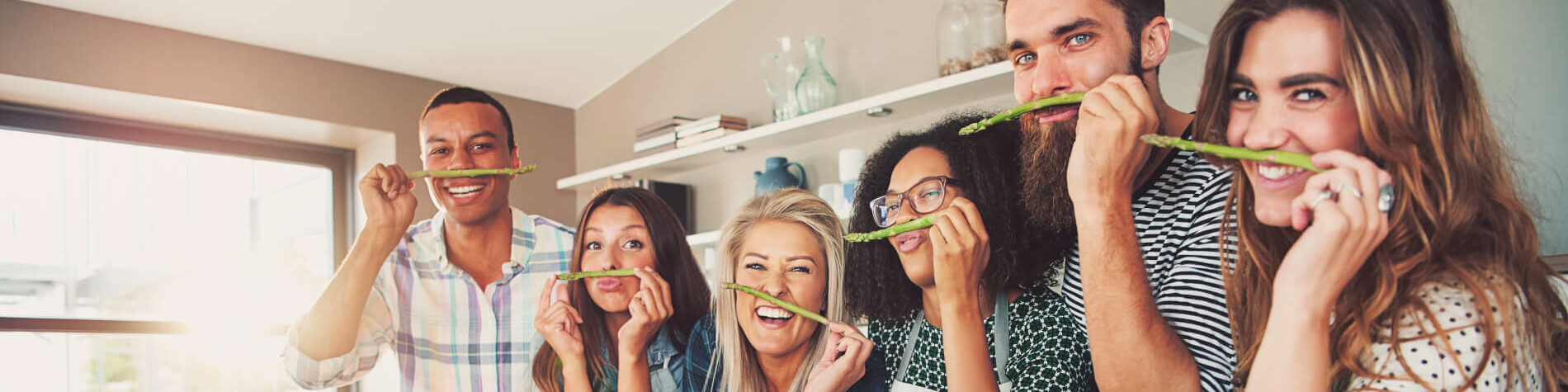 Vier Frauen und zwei Männer vor Töpfen und halten sich grünen Spargel über die Lippe.