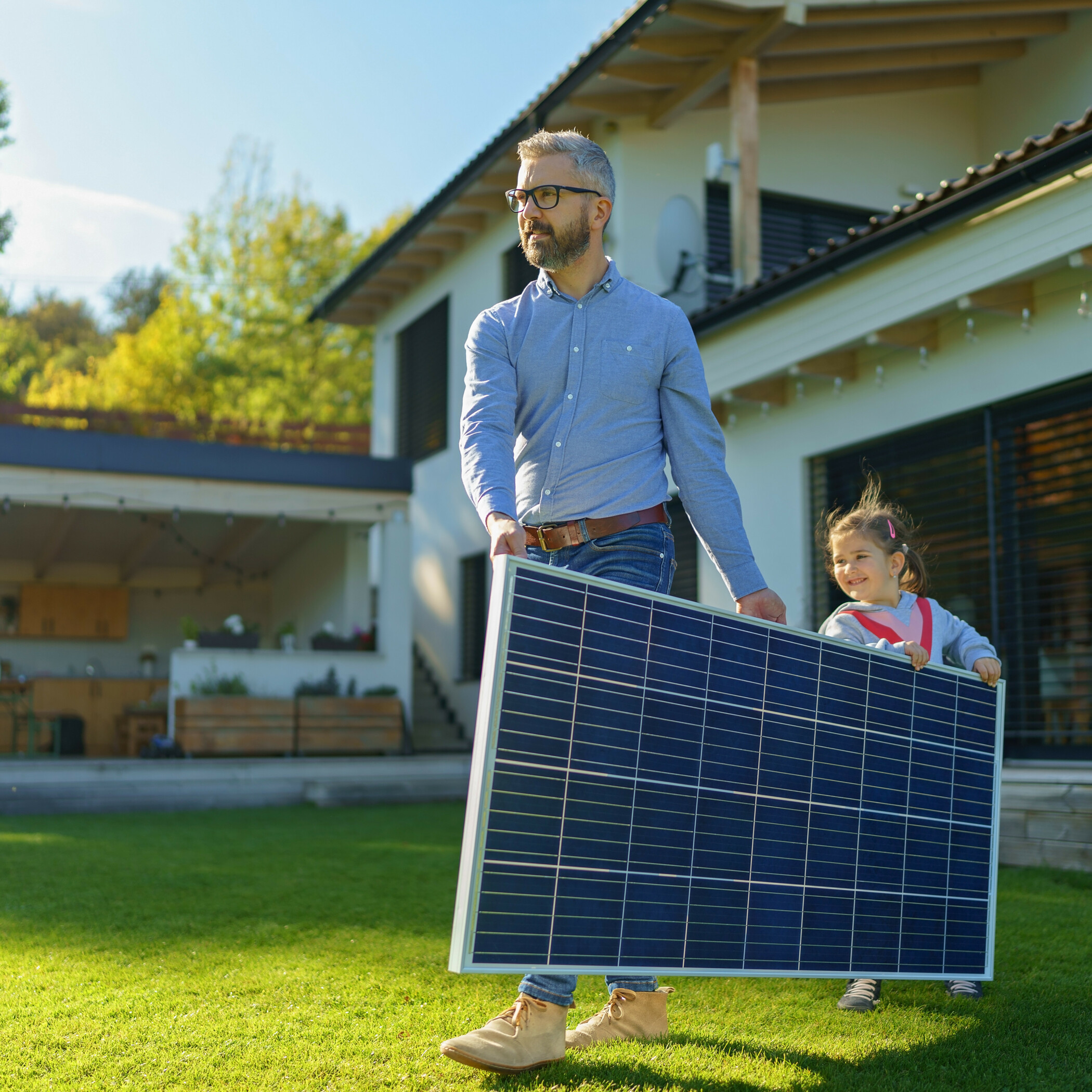 Vater trägt mit seiner Tochter ein Solarmodul durch den Garten.