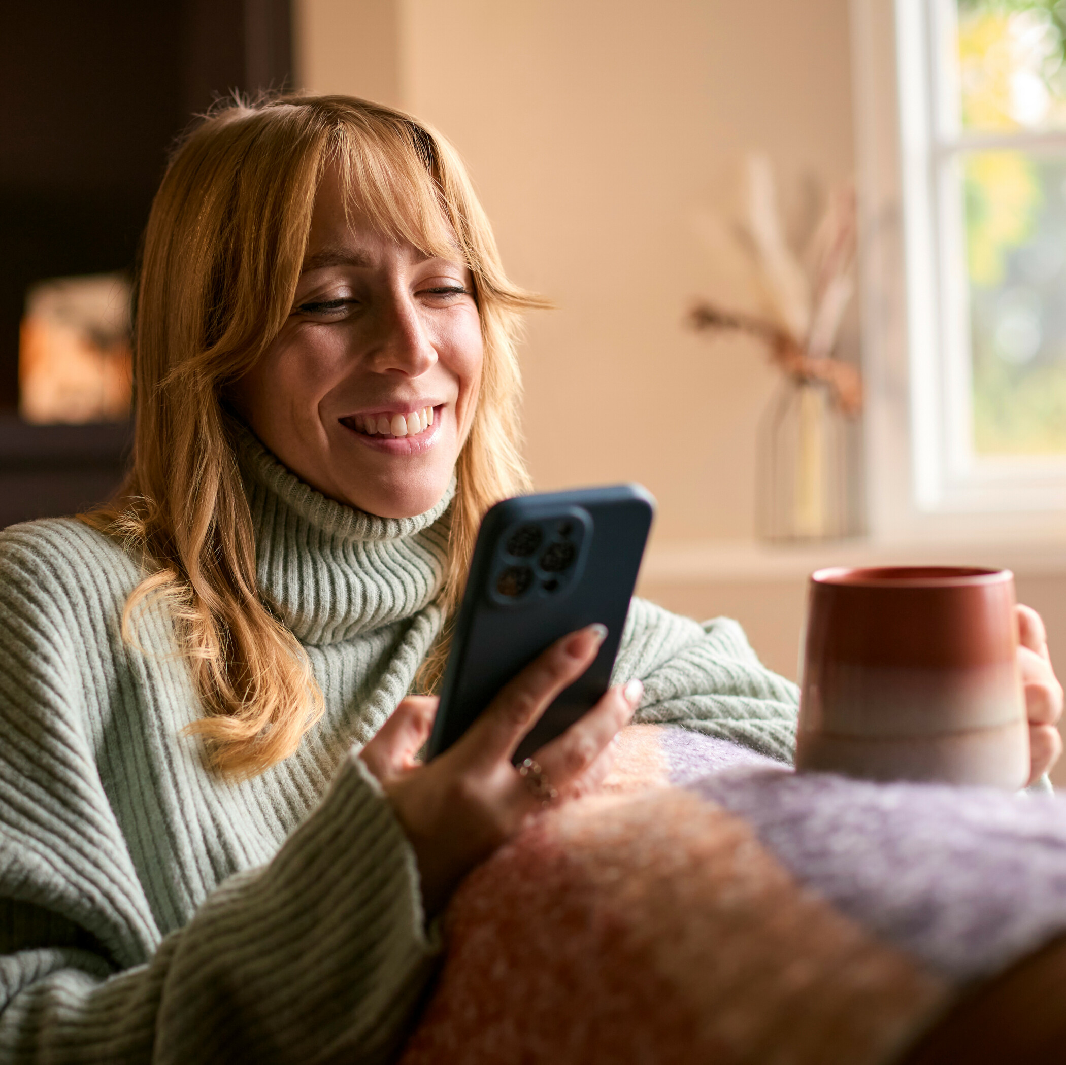 Lächelnde Frau mit grünem Pullover, eine Tasse in der einen und ein Handy in der anderen Hand.