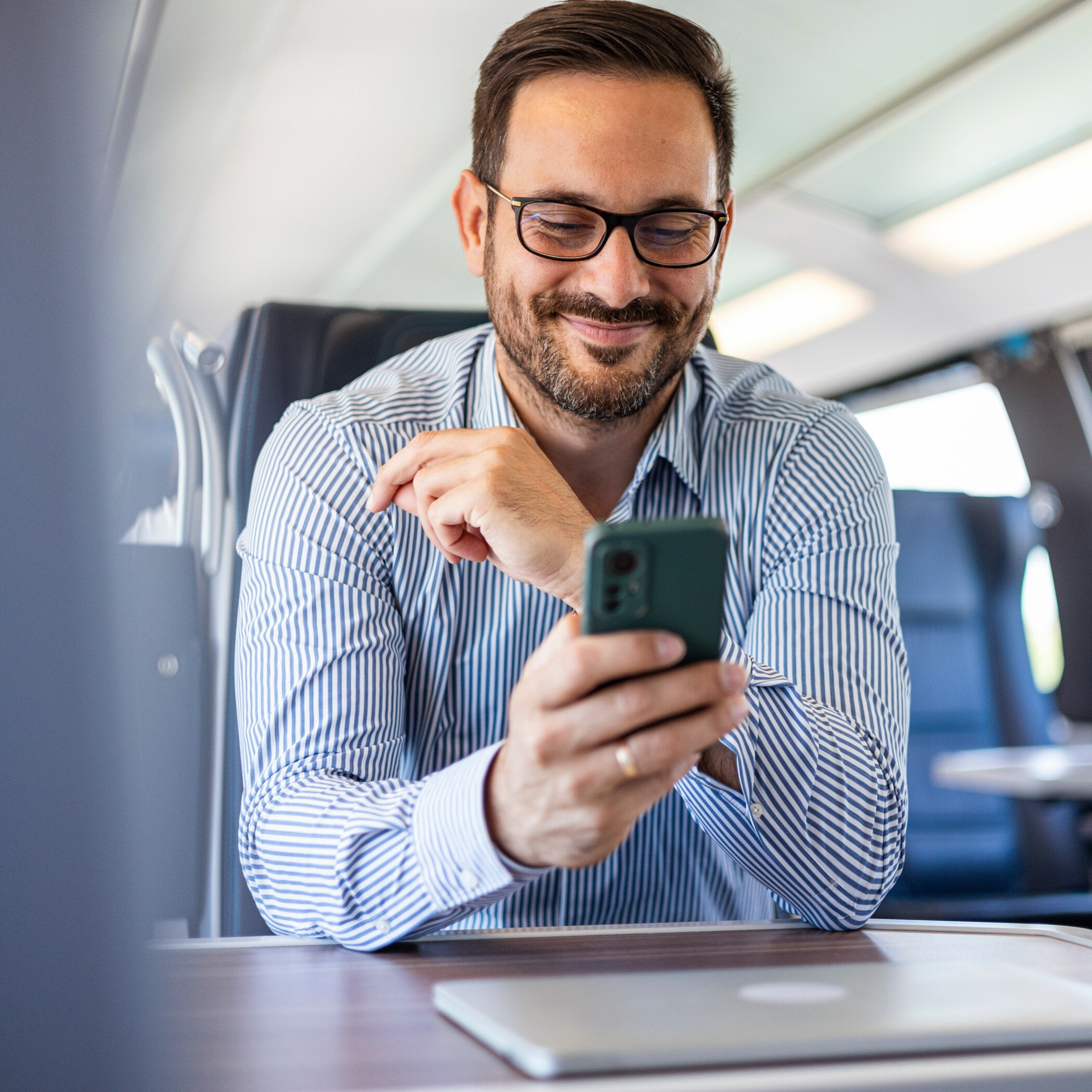 Businessmann mit blau gestreiftem Hemd sitzt im Zug und schaut lächelnd auf sein Handy.