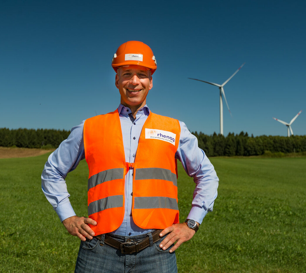 rhenag Mitarbeiter mit orangener Weste und orangenem Helm steht auf einer Grünfläche mit Windrädern im Hintergrund. Er befindet sich im Windpark in Höhn, Westerwald.