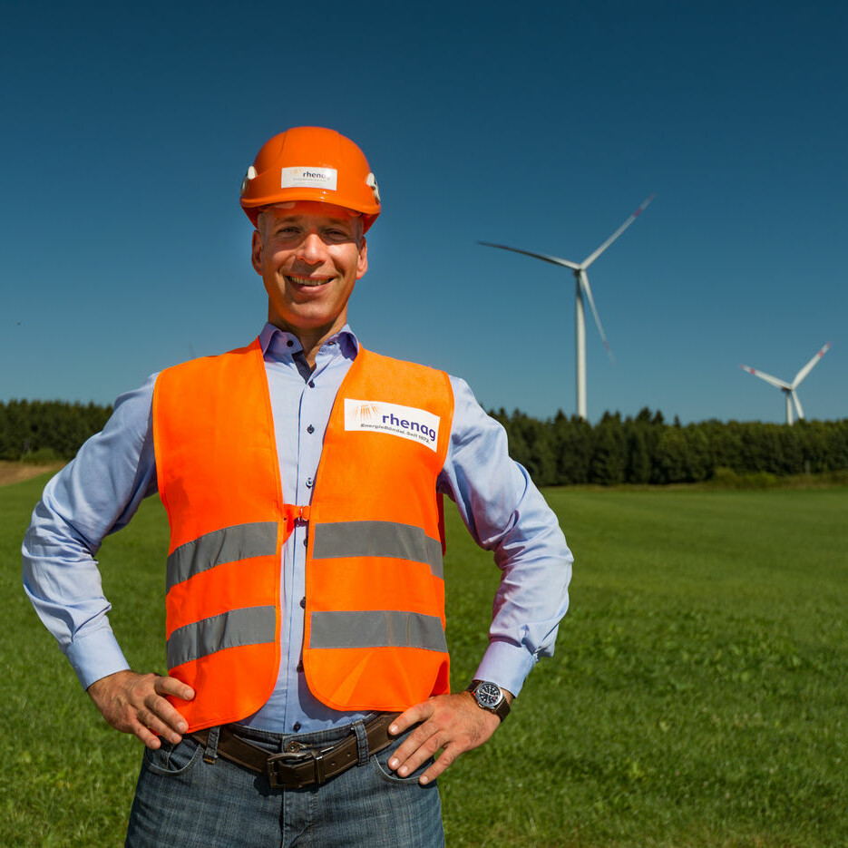 rhenag Mitarbeiter mit orangener Weste und orangenem Helm steht auf einer Grünfläche mit Windrädern im Hintergrund. Er befindet sich im Windpark in Höhn, Westerwald.