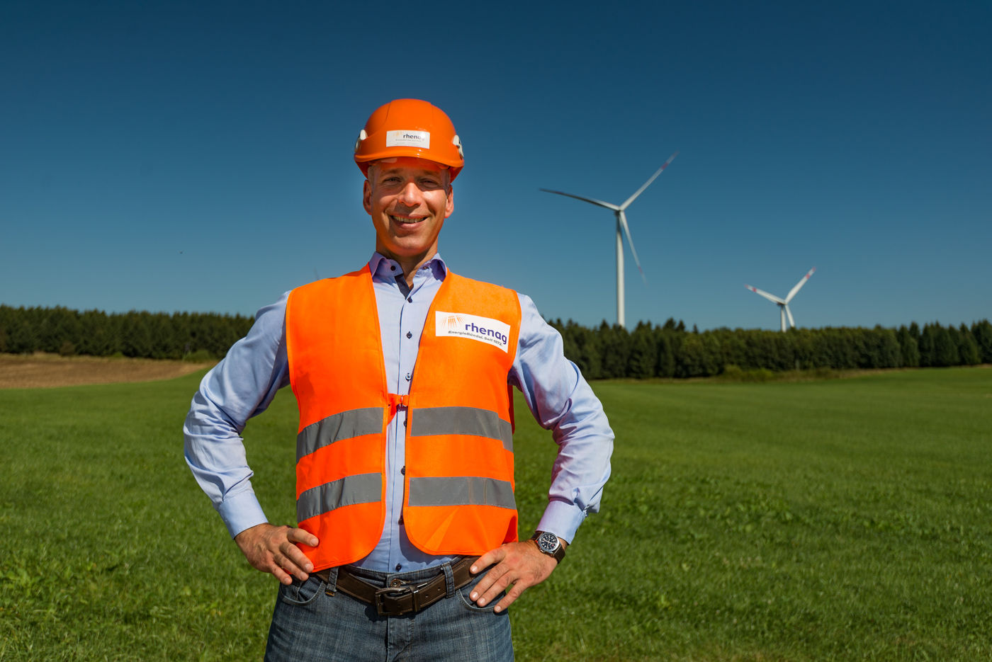 Mann mit orangener Weste und Helm steht auf der Wiese. Im Hintergrund sind zwei Windräder.