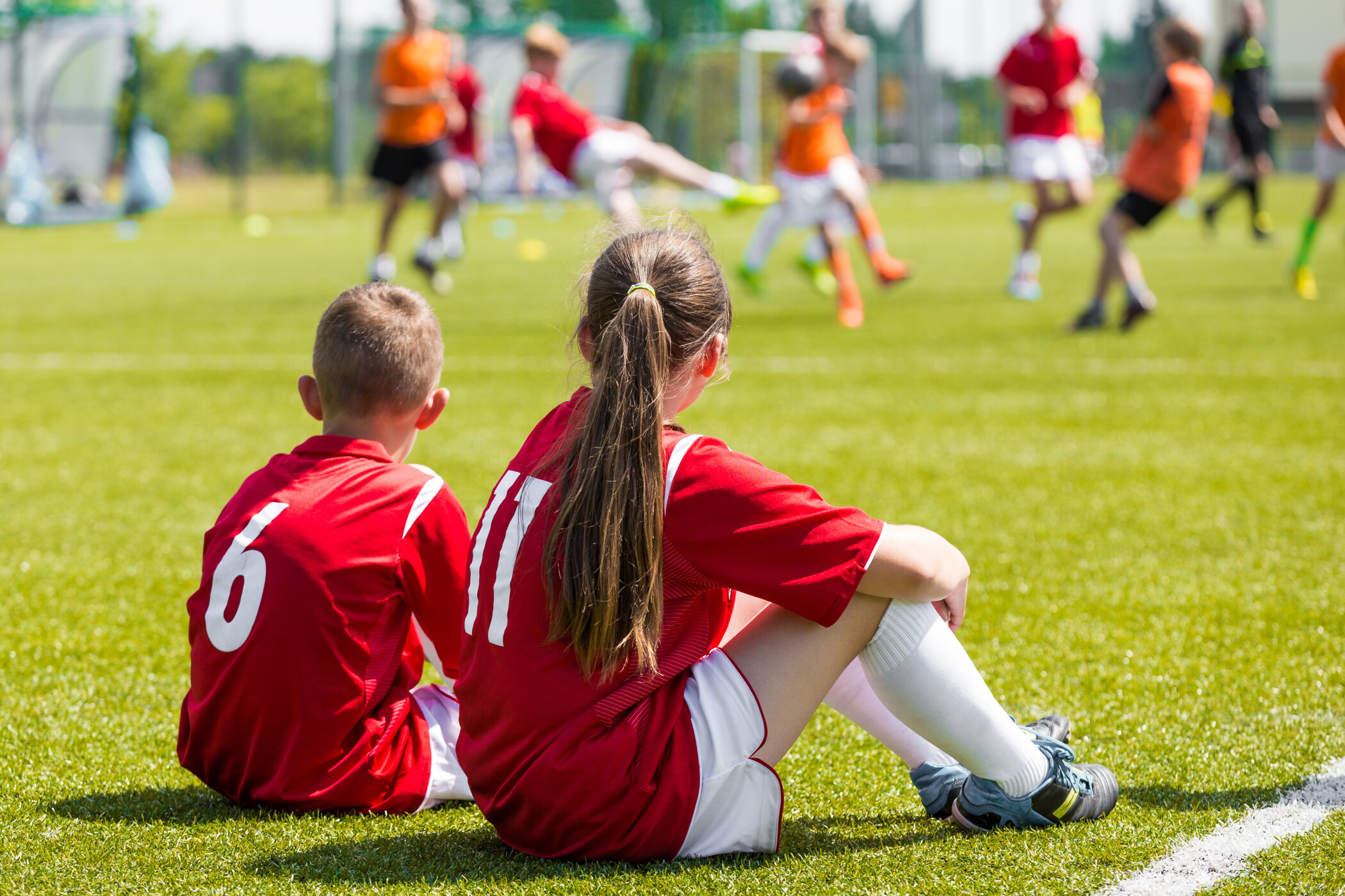 Ein Mädchen und ein Junge sitzen mit dem Rücken zur Kamera auf dem Boden eines Fußballfelds in ihren Trikots und schauen auf die Spieler auf dem Feld.