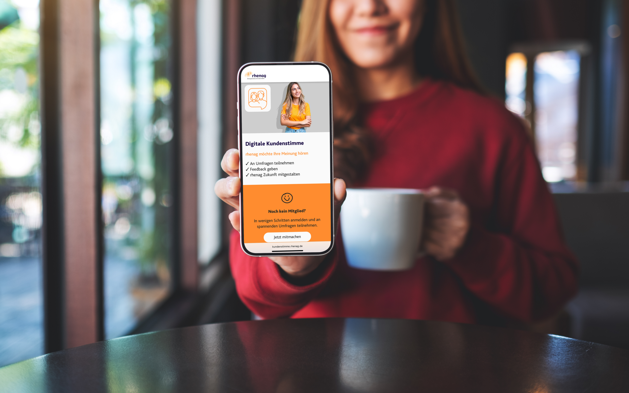 Lächelnde Frau mit rotem Pullover und einer Tasse in der Hand zeigt ihr Handy mit der Website der digitalen Kundenstimme.