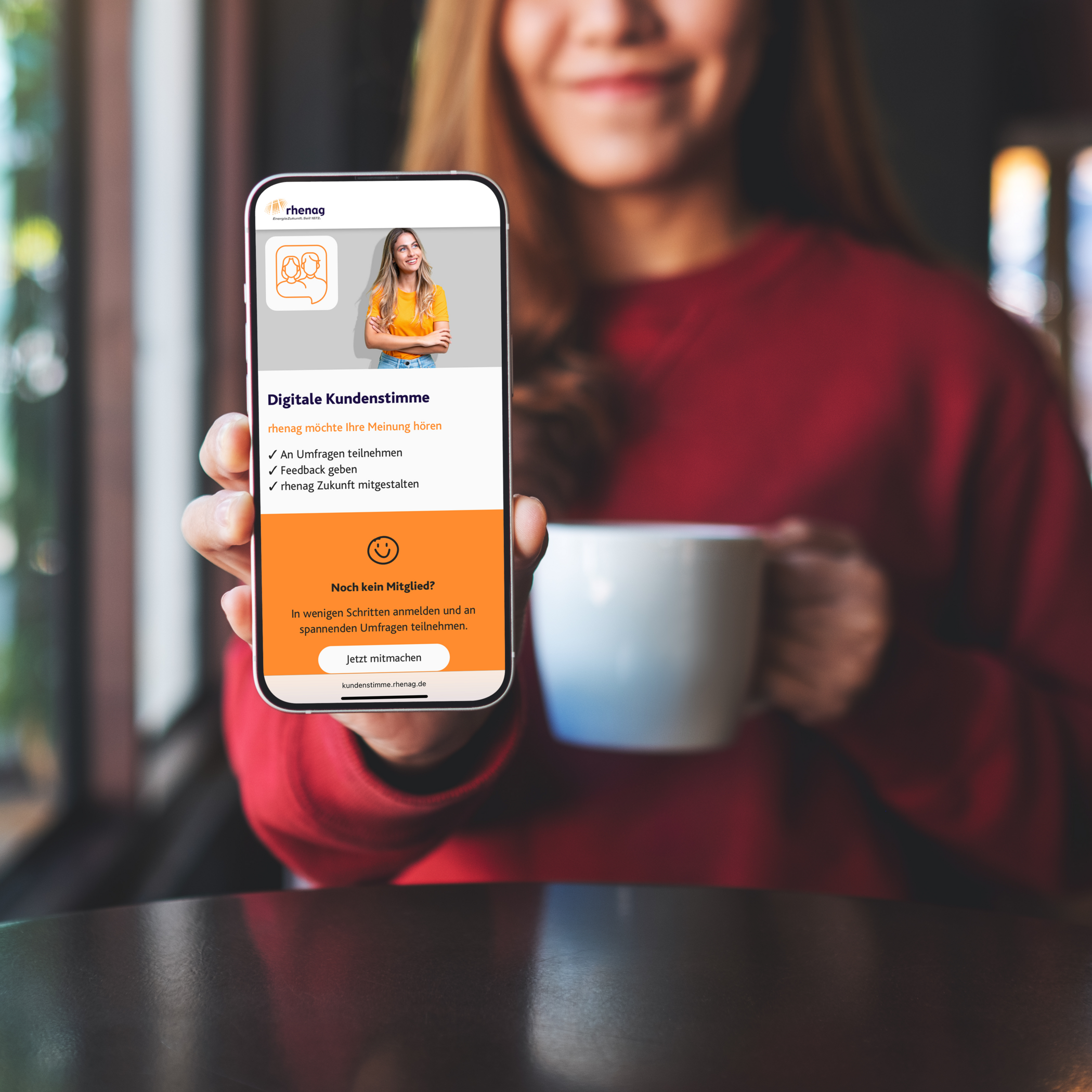 Lächelnde Frau mit rotem Pullover und einer Tasse in der Hand zeigt ihr Handy mit der Website der digitalen Kundenstimme.