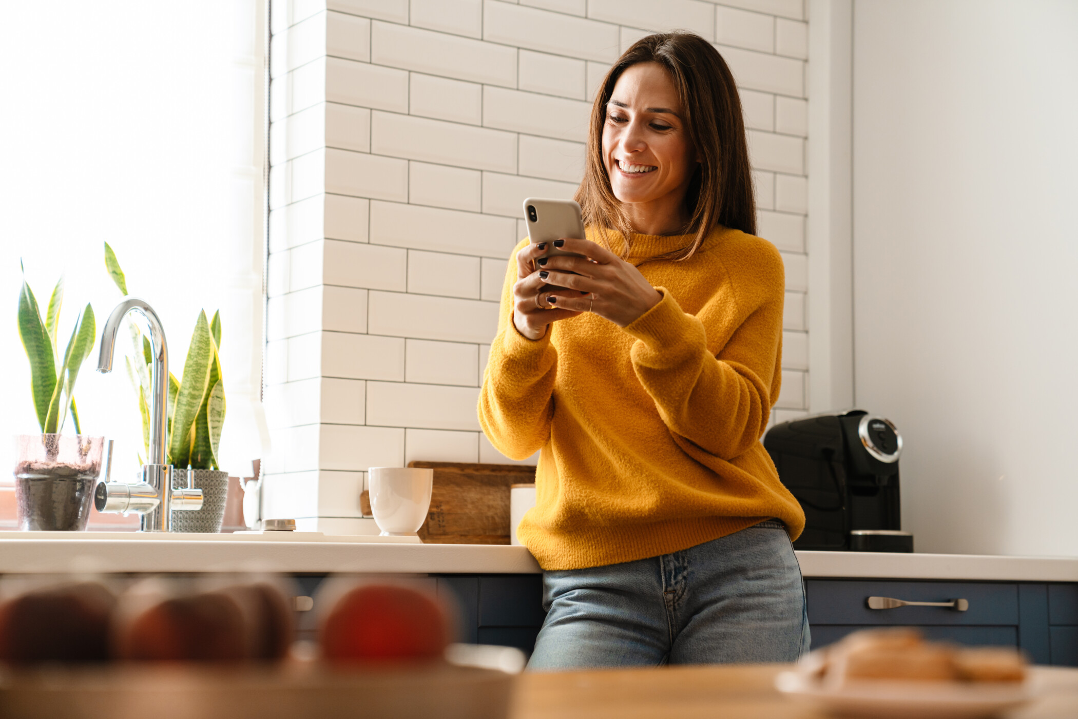 Frau mit gelbem Pullover steht in der Küche und schaut lächelnd auf ihr Handy.