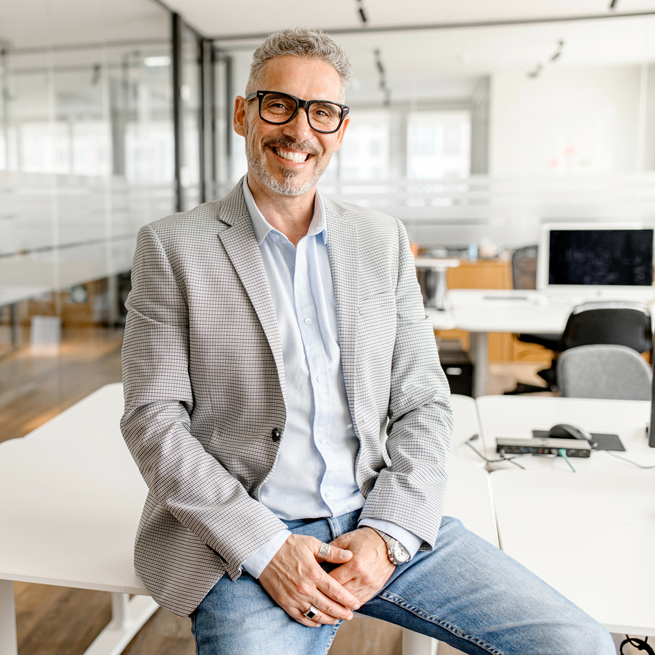 Businessmann mittleren Alters in grauem Jackett sitzt auf einem Tisch im Büro mit Glaswänden im Hintergrund.