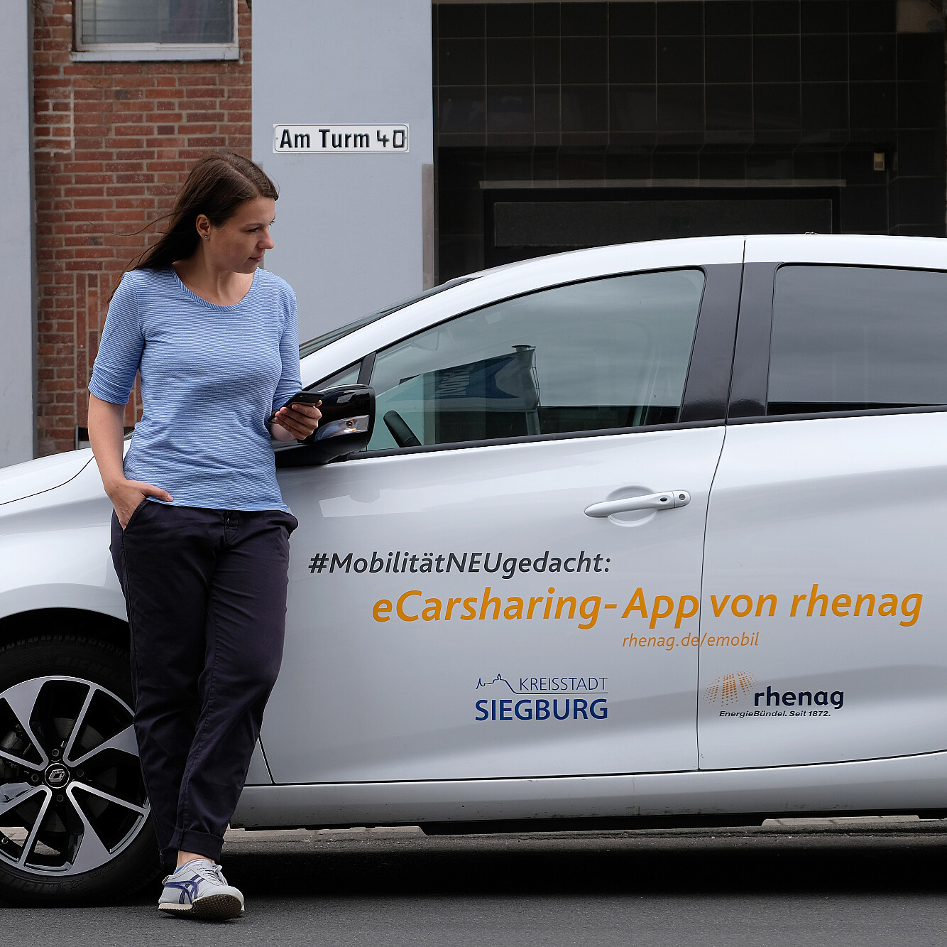 Frau mit Handy in der Hand lehnt am rhenag E-Carsharing-Auto.