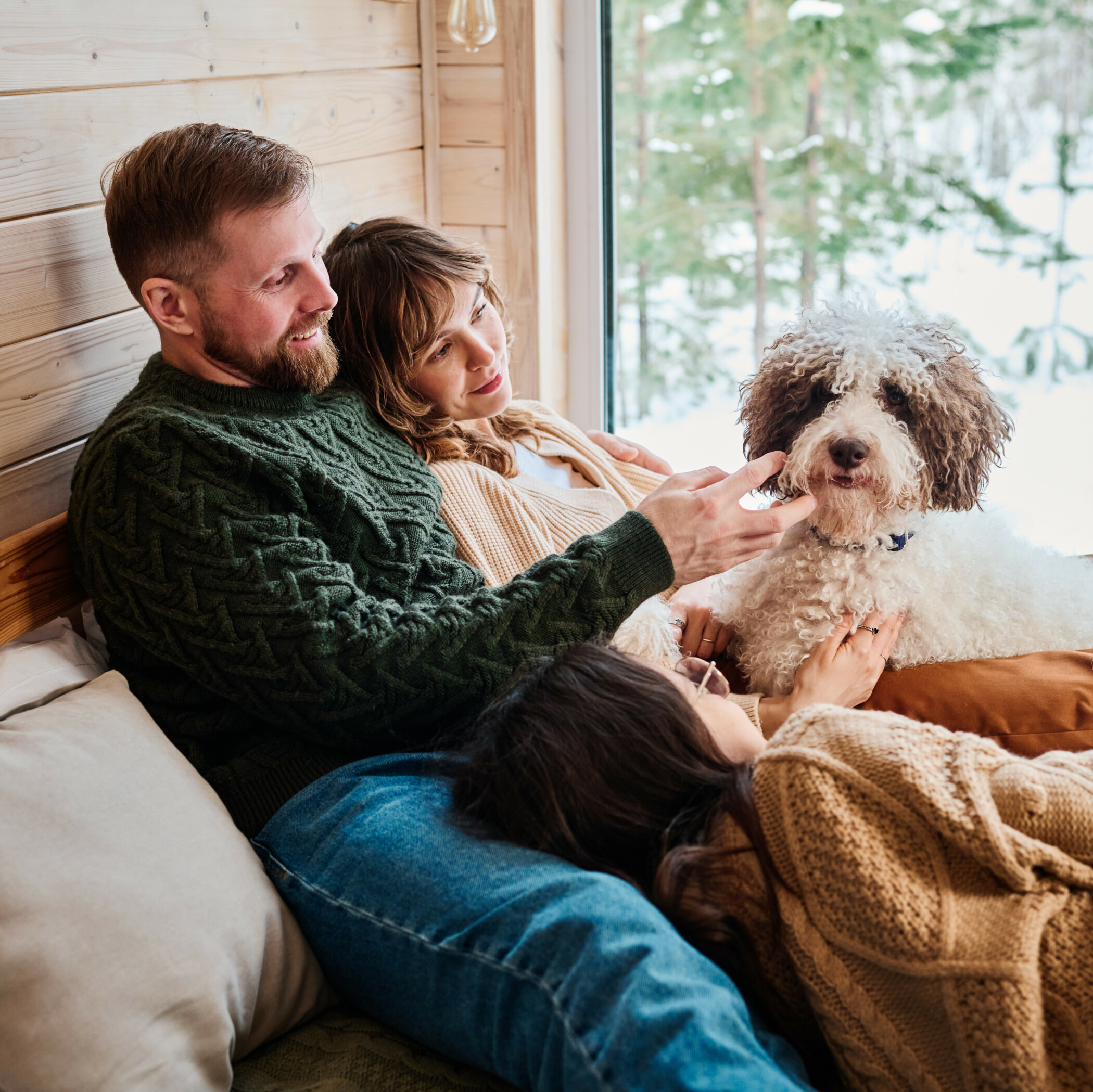 Vater, Mutter und Tochter liegen mit ihrem Hund auf dem Sofa ihres Holzhauses und durch das große Fenster sieht man einen verschneiten Wald.