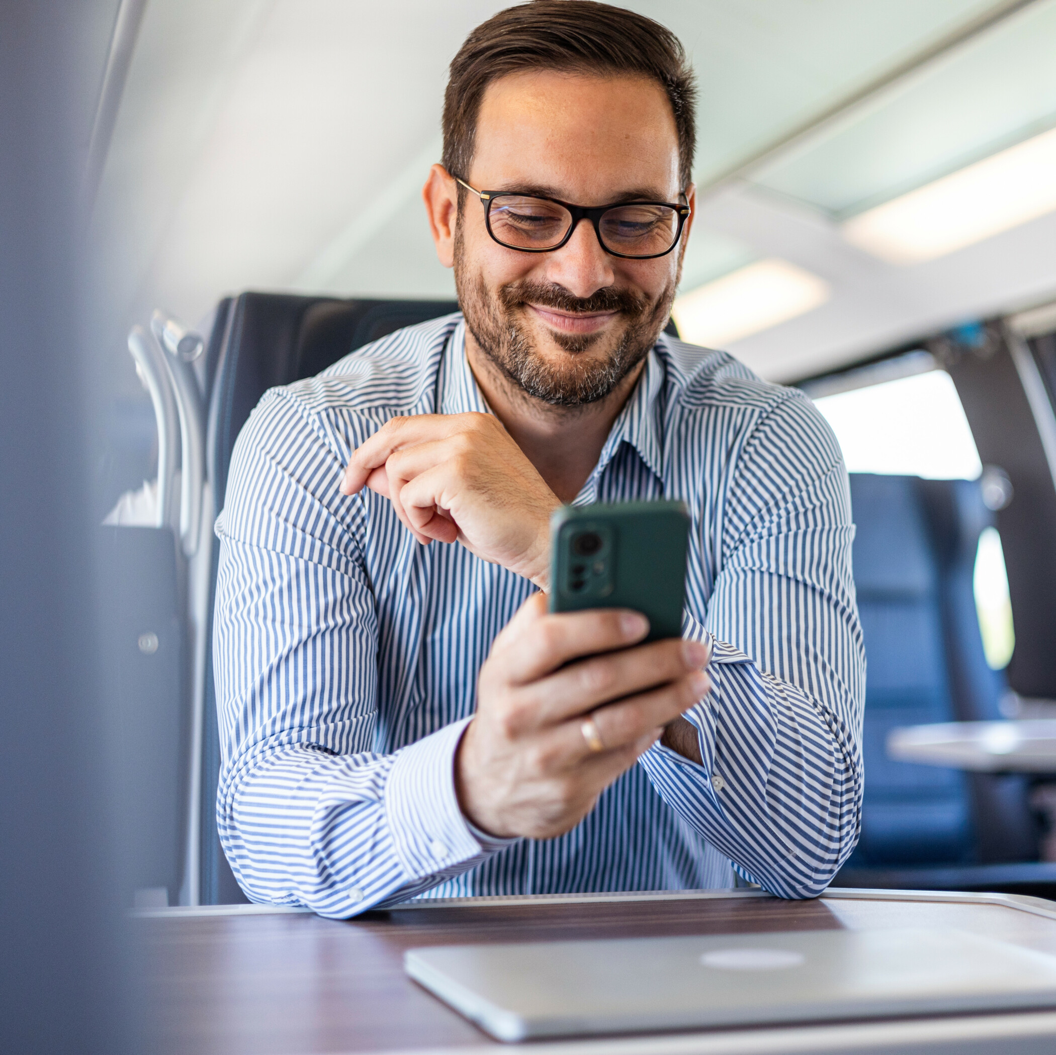 Geschäftsmann im blau-weiß-gestreiftem Hemd sitzt im Zug und schaut auf sein Handy.