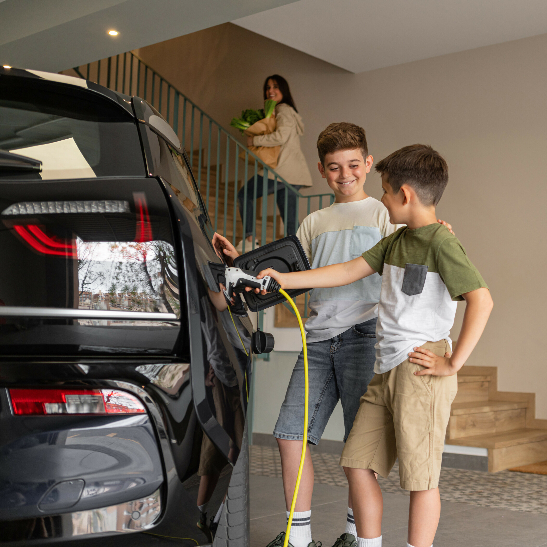 Kinder laden E-Auto Zwei Kinder laden in der Garage das Elektroauto, während die Mutter mit Einkäufen die Treppe hochgeht.