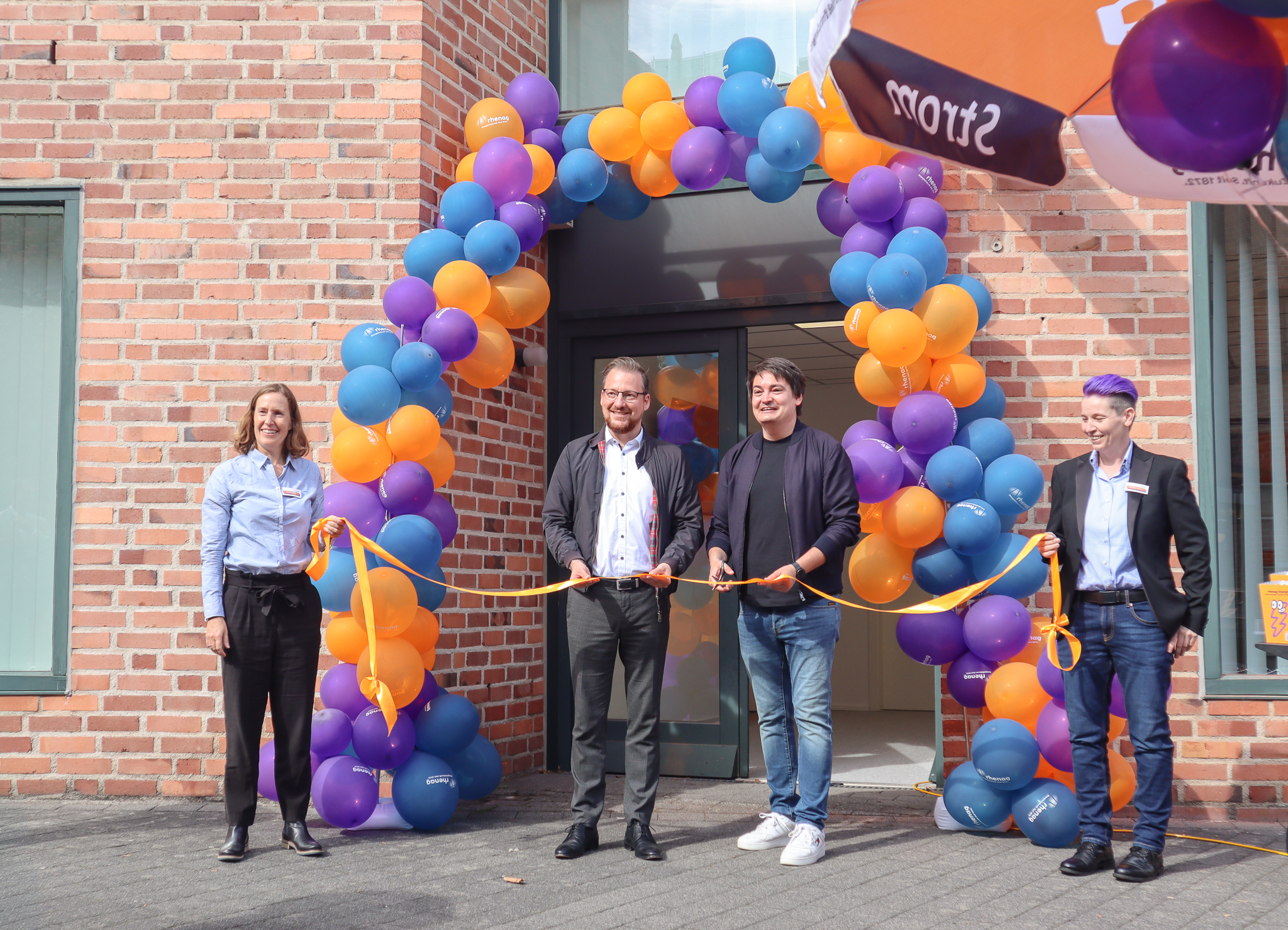 Vier Personen stehen vor dem neuen rhenag Kunden-Center in Hennef. Zwei von ihnen durchschneiden ein orangefarbenes Band am Eingang, der mit bunten Luftballons geschmückt ist.