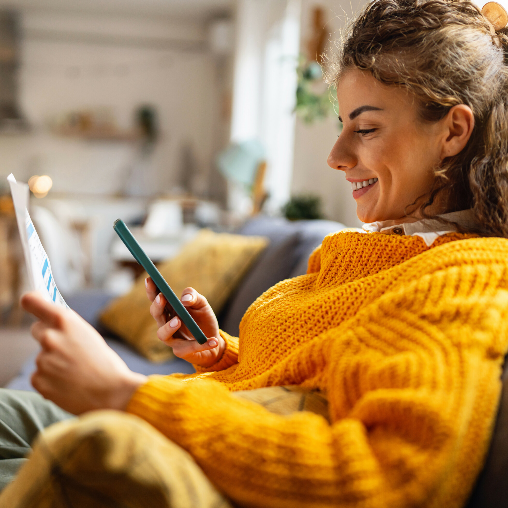 Lächelnde Frau im gelben Pulli sitzt mit einem Formular auf der Couch und schaut gleichzeitig auf ihr Handy.