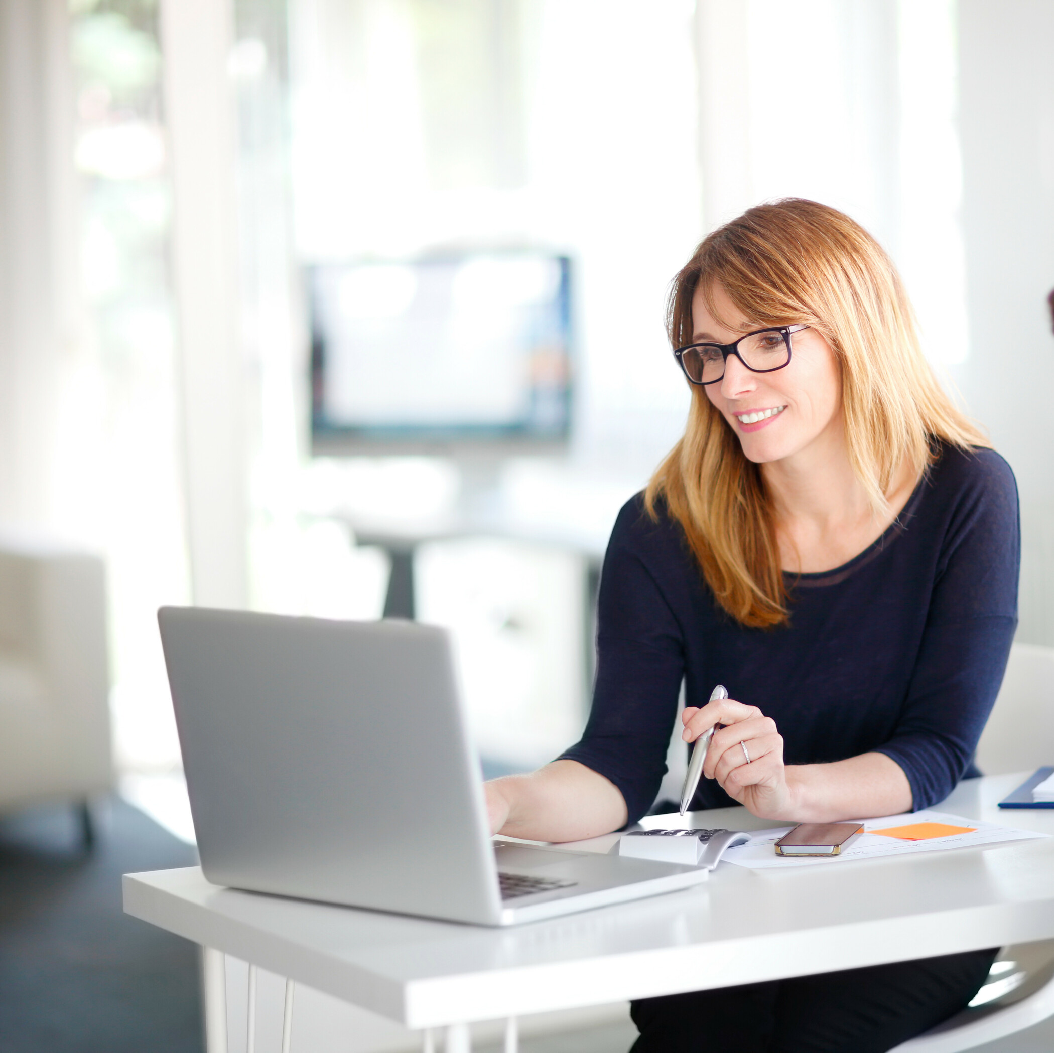 Frau mit Kugelschreiber in der Hand sitzt am Laptop an einem weißen Tisch.