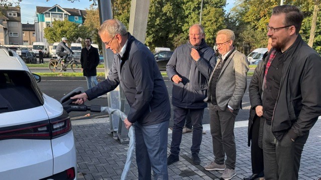 Eine Person lädt ein Elektrofahrzeug an einer Schnellladesäule, während mehrere weitere Personen den Ladevorgang an einem überdachten Ladepark beobachten